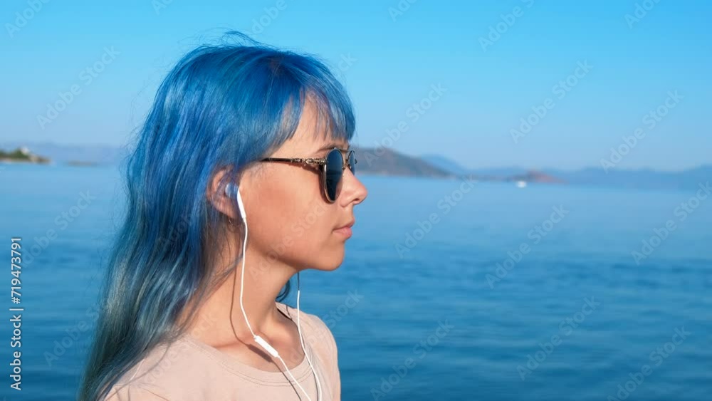 Contemporary girl in headphones by water. A nice girl with blue hair listen the music in earphones and look into the river distance in summer. Contemporary girl in headphones by water. A nice girl with blue hair listen the music in earphones and look into the river distance in summer.