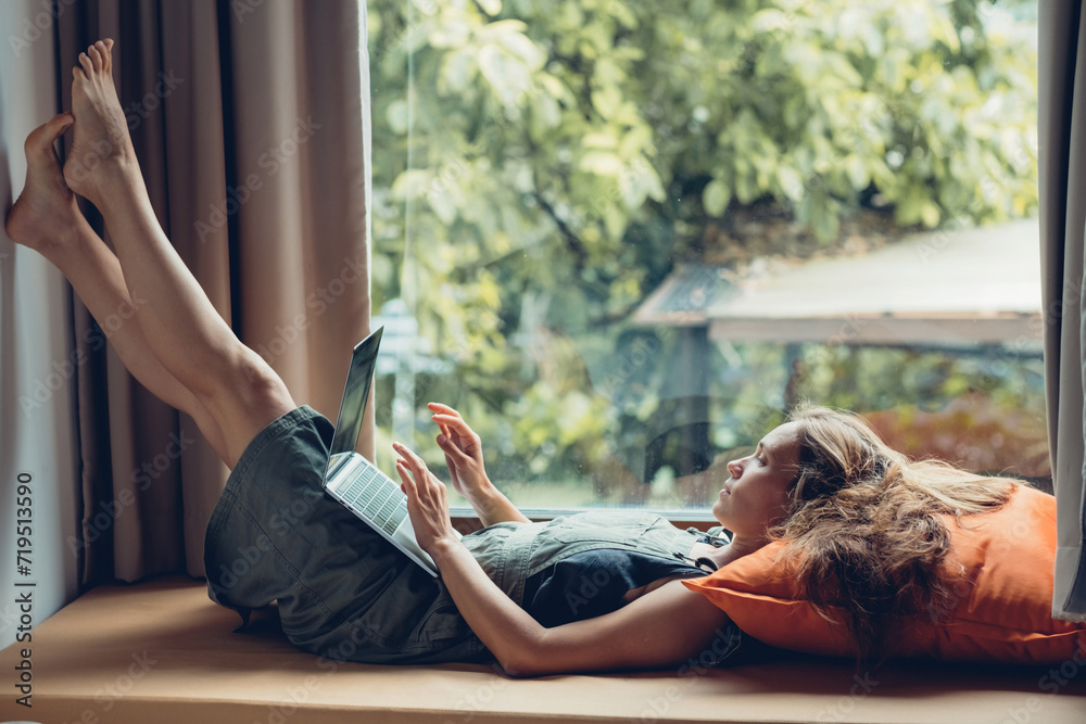Young woman working on laptop while lying on windowsill at home. Work from home in cozy atmosphere.