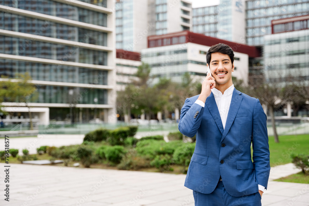 A Hispanic businessman is engaging in a conversation on his mobile phone, smiling subtly with an air of success as he stands before an urban landscape, embodying the modern multitasking professional. A Hispanic businessman is engaging in a conversation on his mobile phone, smiling subtly with an air of success as he stands before an urban landscape, embodying the modern multitasking professional.