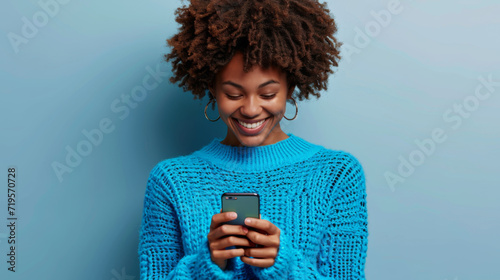 young woman with a vibrant afro hairstyle is smiling and looking at her smartphone, wearing a cozy turquoise turtleneck sweater against a matching turquoise background