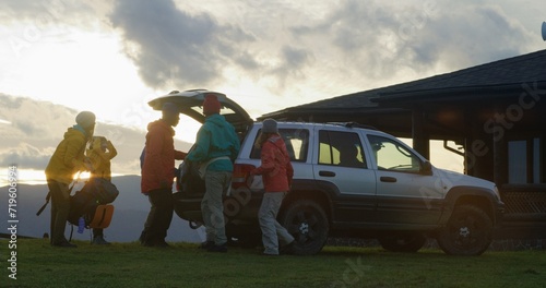 Full shot of diverse tourist friends arrive to rental house on hill. Happy hikers take luggage from trunk of car and talk. Hiking buddies during trip or expedition to mountains on their vacation.