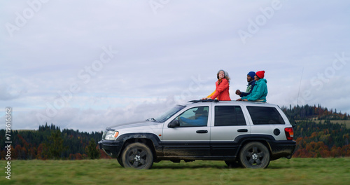 Group of diverse tourists together ride on vacation in mountains. Hikers standing up through sunroof of car sing, dance and wave hands on road trip. People enjoy drive in car hatch. Active recreation.