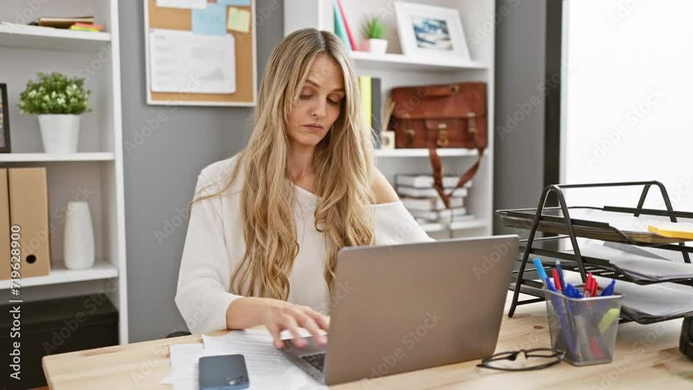 A focused blonde woman working with smartphone and laptop in a modern office setting A focused blonde woman working with smartphone and laptop in a modern office setting