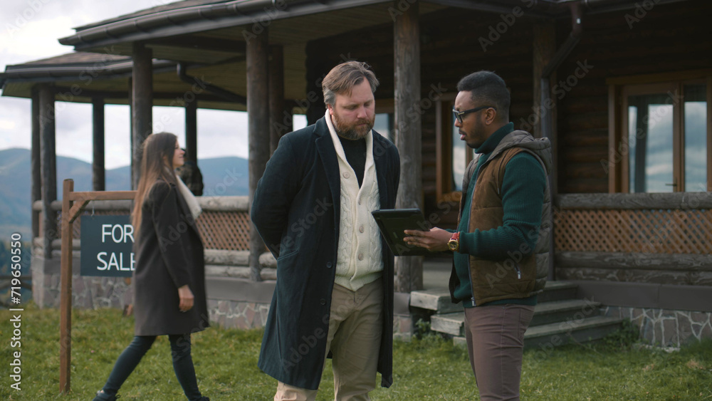 Family with kids visit potential home property. African American real state agent consults Caucasian man and shows plan of modern house on digital tablet computer. Residential property on sale concept