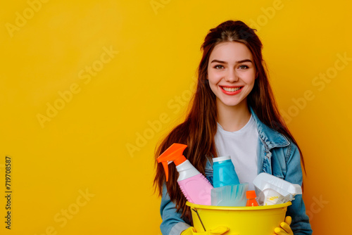 Smiling young woman in rubber protective gloves holding bucket with cleaning supplies on yellow background. Spring cleaning, housekeeping and hygiene concept. Design for banner, poster with copy space