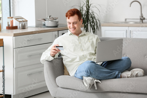 Handsome young man with credit card and modern laptop sitting on sofa while shopping online