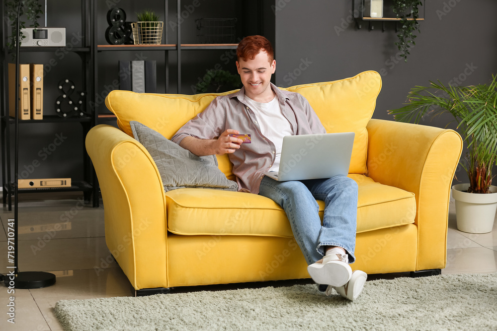 Handsome young man with credit card and modern laptop sitting on sofa while shopping online