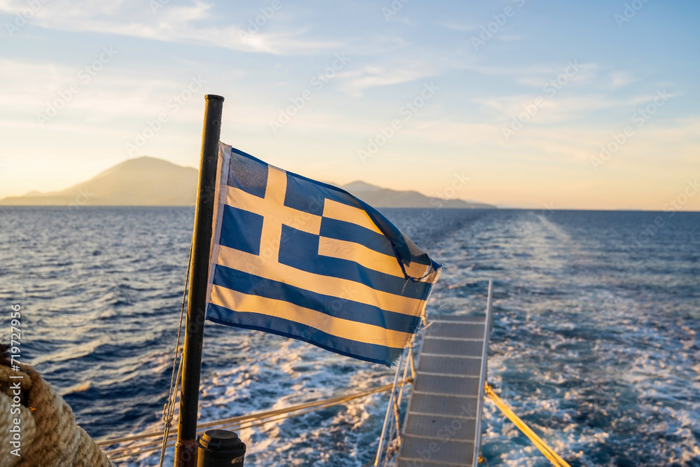 Sunset over the sea with the Greek flag in the foreground viewed from the ferry on the route to Zakynthos island in Geece