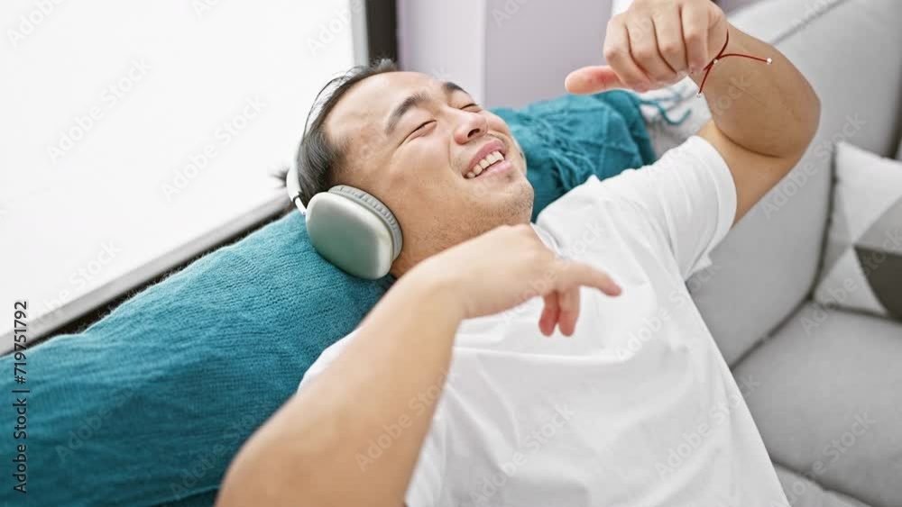 Happy chinese guy dancing joyfully, in-home fun while he's listening to music, sitting on his living room sofa with earphones