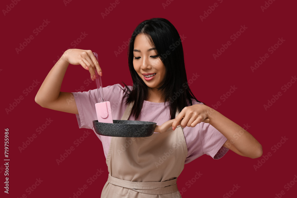 Portrait of young Asian woman with frying pan and spatula on red background Portrait of young Asian woman with frying pan and spatula on red background