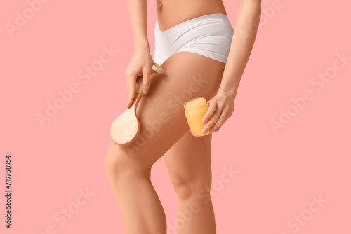 Beautiful young woman with cellulite problem, massage brush and jar of body scrub on pink background