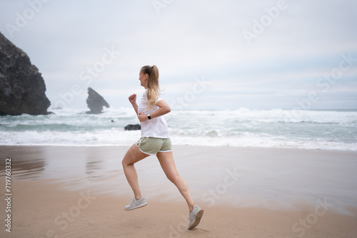 Full length of athletic woman running along seashre. Young sportswoman in white top and sporty shorts jogging on sandy beach. Fit lady represents healthy lifestyle.