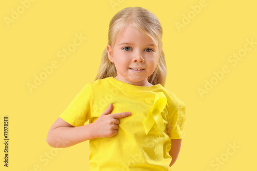 Cute little girl pointing at yellow ribbon on color background. Childhood cancer awareness concept