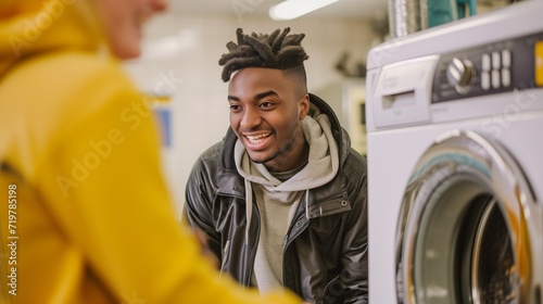 Young black man with leather jacket and grey hoodie, handsome smiling face, waiting a friend doing laundry in a launderette, the two men are using a washing machine to clean their clothes and talking photo