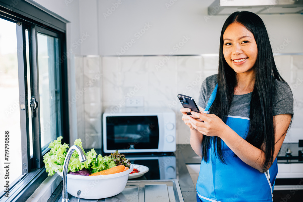 Smiling woman leans on the kitchen table reading messages on her cellphone while cooking dinner. Illustrating a cheerful housewife's modern multitasking routine at home. mobile phone Smiling woman leans on the kitchen table reading messages on her cellphone while cooking dinner. Illustrating a cheerful housewife's modern multitasking routine at home. mobile phone