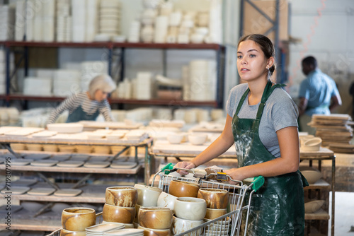 Young woman in ceramics factory carries many different clay ceramic cups and plates in a cart