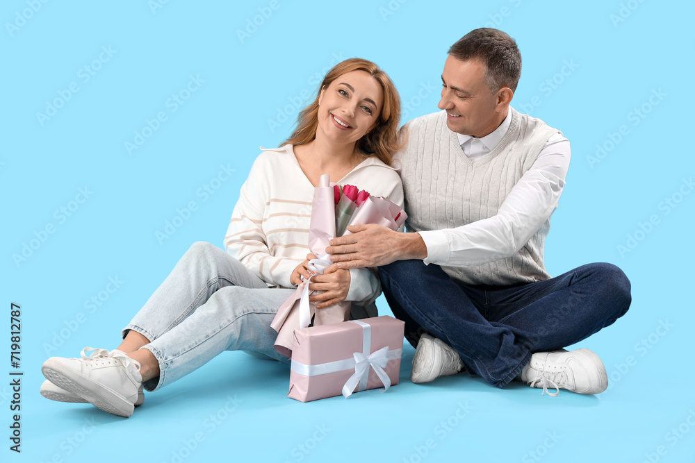 Middle-aged couple with bouquet of flowers and gift box sitting on blue background. International Women's Day Middle-aged couple with bouquet of flowers and gift box sitting on blue background. International Women's Day