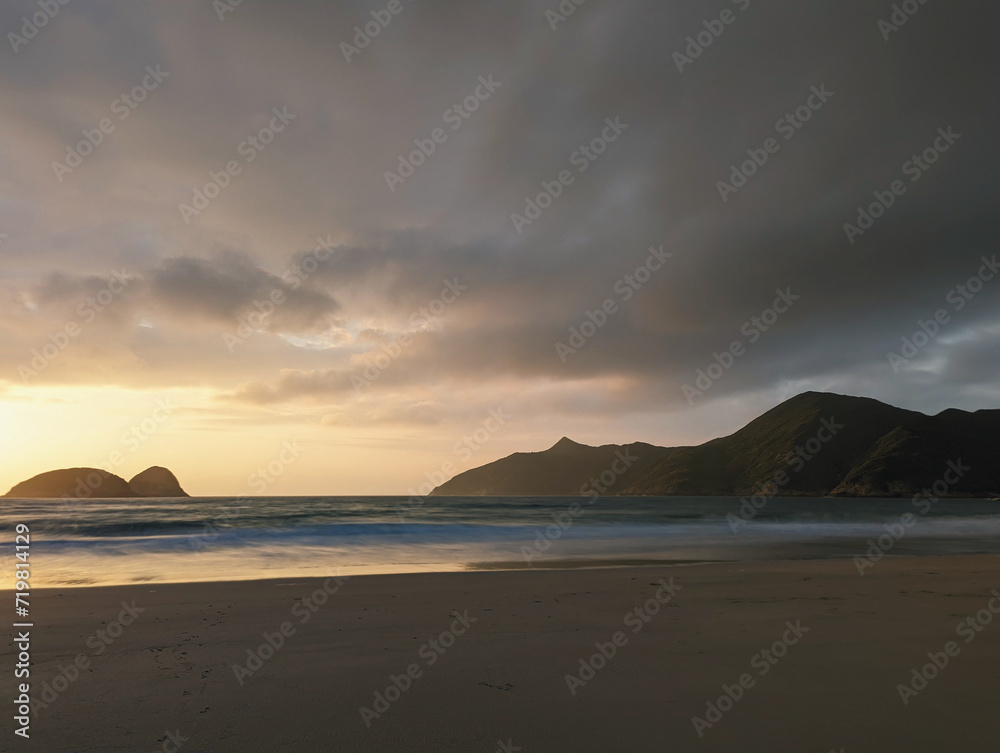 A serene beach at dusk, with gentle waves lapping the shore and dark clouds overhead, illuminated by the fading sunlight.