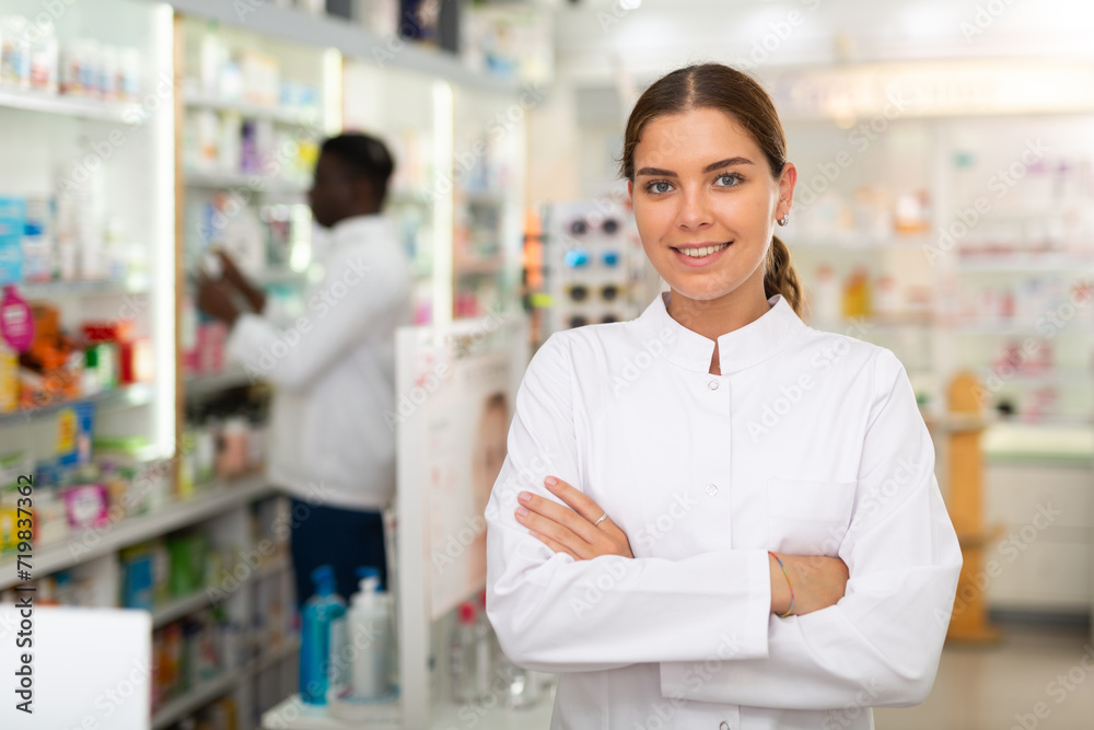 Portrait of female pharmacist standing in salesroom of drugstore and demonstrating haircare product. Her colleague working behind.