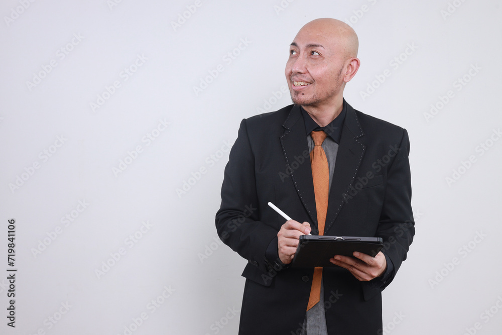 Happy middle aged Asian business man ceo wearing suit standing in office using digital tablet and looking away Happy middle aged Asian business man ceo wearing suit standing in office using digital tablet and looking away
