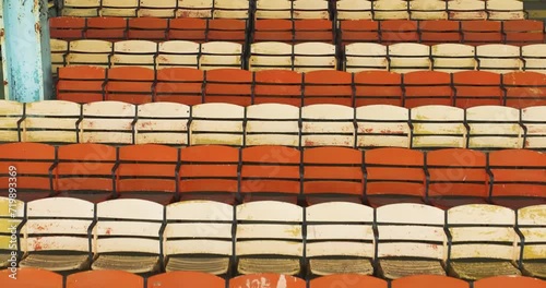 Drone, aerial, video of old faded orange and yellow painted wooden seats at a abandoned a stadium grandstand.
