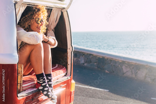 Happy adult beautiful woman enjoy travel lifestyle or summer holiday vacation with old vintage van transport sit down on the back and looking the road and ocean - people free to move and live photo