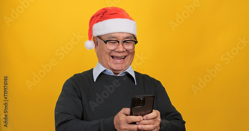 Asian old man wearing a Santa hat and using smart phone. Isolated on yellow background in the studio. © ronnarong