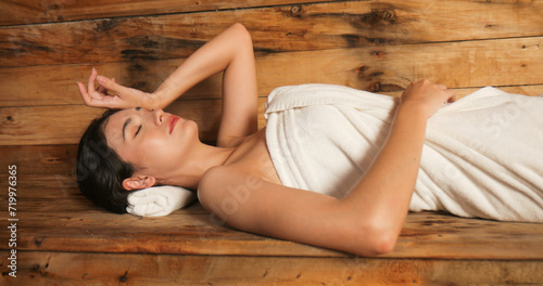 Young woman in towel relaxing in wooden sauna at spa. Asian woman in bathrobe doing body treatment in sauna. © ronnarong