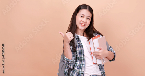 Cute school girl is carrying a school bag and holding a textbook. Isolated on brown background in studio.