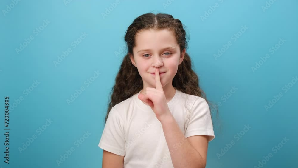 Portrait of adorable funny little girl child making silence gesture with finger on lips, looking away with smile, wearing white t-shirt, posing isolated over blue color background wall in studio Portrait of adorable funny little girl child making silence gesture with finger on lips, looking away with smile, wearing white t-shirt, posing isolated over blue color background wall in studio