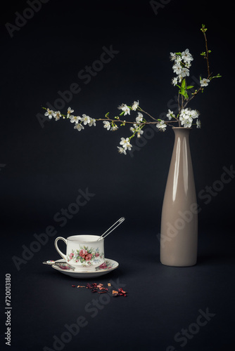 Still life with a beautiful fine china tea cup, tea accessories and a blossoming twig in a vase on a black background