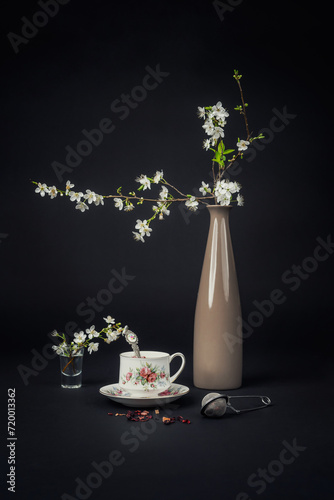 Still life with a beautiful fine china tea cup, tea accessories and a blossoming twig in a vase on a black background