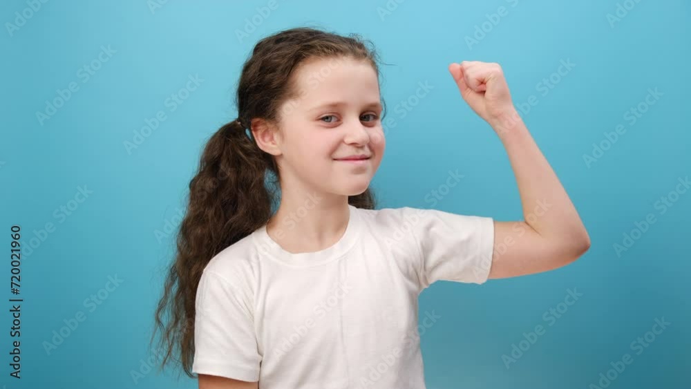 Portrait of smiling little girl child pointing at biceps, feeling powerful and self-confident, showing strength, wearing white t-shirt, posing isolated over blue color background wall in studio Portrait of smiling little girl child pointing at biceps, feeling powerful and self-confident, showing strength, wearing white t-shirt, posing isolated over blue color background wall in studio