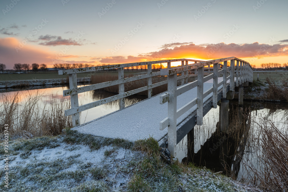 White wooden arched bridge with a layer of snow in the morning sun. Behind Lake Dirkshorn lies this picturesque white arched bridge, as part of a walking route through the beautiful North Holland land