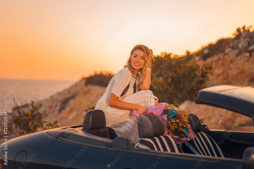 Happy beautiful blonde in a white boho style dress with a beautiful bouquet of flowers in a black convertible against the backdrop of the sea sunset light.