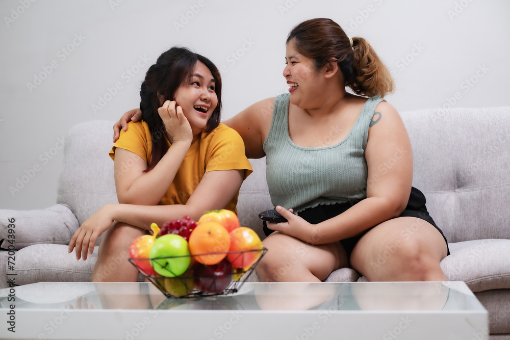 Two young overweight women sitting on sofa in living room looking each other with smile and laugh, exciting emotion at home. Two young overweight women sitting on sofa in living room looking each other with smile and laugh, exciting emotion at home.