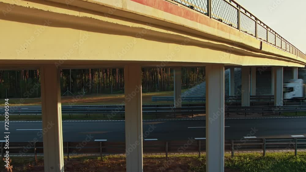 Empty and loaded flatbed trailers trucks are driving on highway under overpass in countryside. Vehicles move on roadway under flyover overbridge. Traffic barrier guard rail on four-lane roadway Empty and loaded flatbed trailers trucks are driving on highway under overpass in countryside. Vehicles move on roadway under flyover overbridge. Traffic barrier guard rail on four-lane roadway