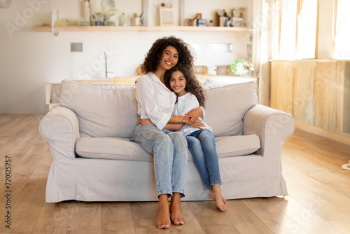 Loving young mother hugging her daughter sitting together on sofa at home in living room interior. Mother's love, motherhood concept