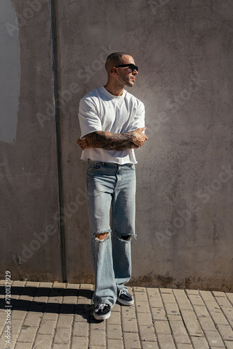 Chico joven musculado y tatuado posando con ropa urbana en la calle con luz dura del sol