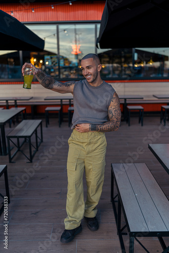 Chico joven musculado y tatuado posando con ropa urbana en terraza de restaurante de comida rapida bebiendo un refresco de naranja 
