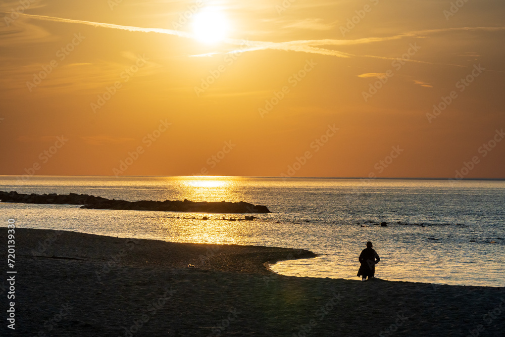 A woman in silhouette walks on a sandy beach as the A new day begins: woman in silhouette  walks on beach  as sun rises over lake ontario shot on toronto's kew beach room for text
