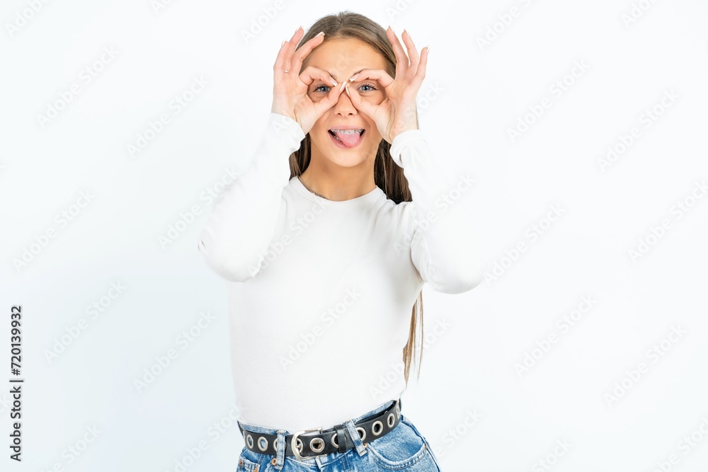 young beautiful caucasian woman wearing white long sleeve T-Shirt doing ok gesture like binoculars sticking tongue out, eyes looking through fingers. Crazy expression. young beautiful caucasian woman wearing white long sleeve T-Shirt doing ok gesture like binoculars sticking tongue out, eyes looking through fingers. Crazy expression.