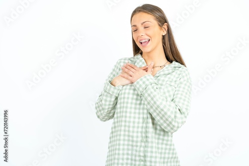 Beautiful young woman wearing green plaid pyjama expresses happiness, laughs pleasantly, keeps hands on heart