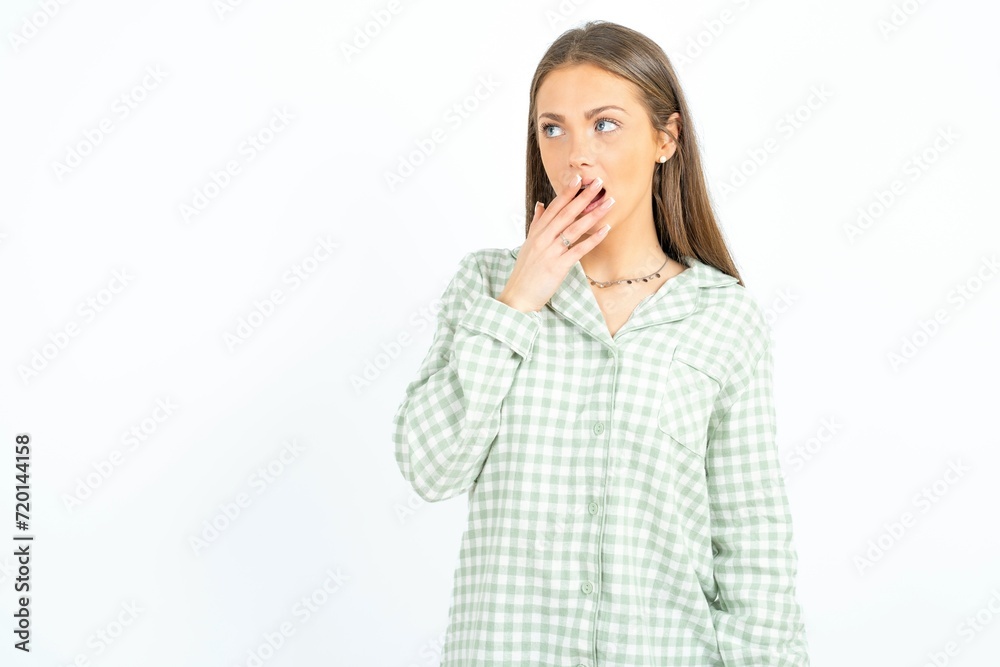 Beautiful young woman wearing green plaid pyjama covers mouth and looks with wonder at camera, cannot believe unexpected rumors. Beautiful young woman wearing green plaid pyjama covers mouth and looks with wonder at camera, cannot believe unexpected rumors.