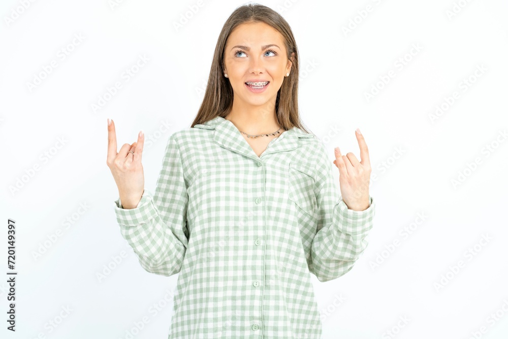 Beautiful young woman wearing green plaid pyjama makes rock n roll sign looks self confident and cheerful enjoys cool music at party. Body language concept. Beautiful young woman wearing green plaid pyjama makes rock n roll sign looks self confident and cheerful enjoys cool music at party. Body language concept.