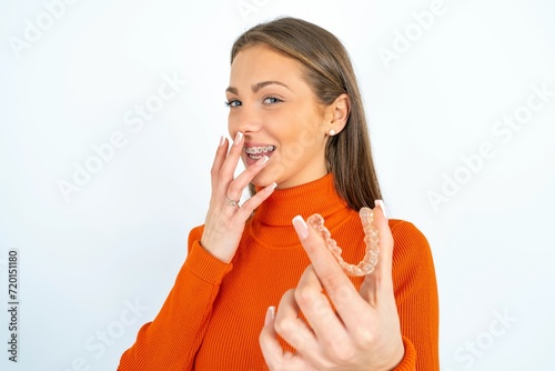 Happy young beautiful caucasian woman wearing orange turtleneck holding and showing at camera an invisible aligner while laughing. Dental healthcare and confidence concept.