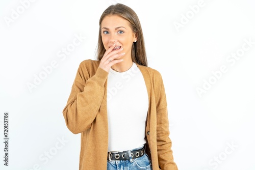 Young beautiful woman standing over white studio background covers mouth and looks with wonder at camera  cannot believe unexpected rumors.