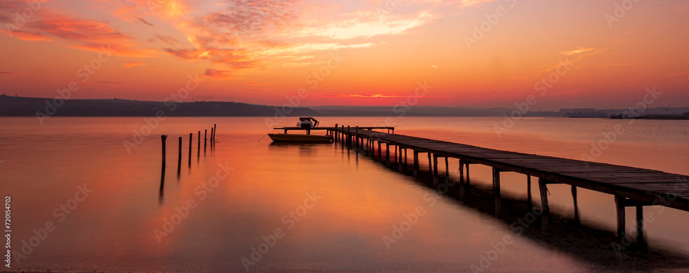 Fototapeta premium Scenic view of exciting red sunset at the shore with a wooden pier and moored boat