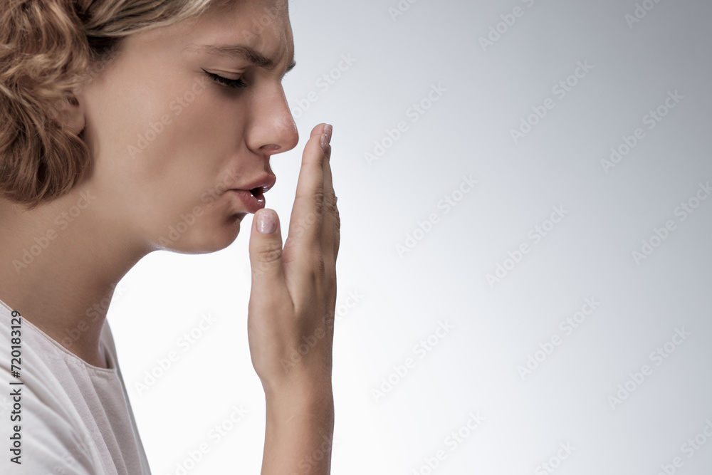 Bad breath. Young girl checking her breath with her hand, blowing to it, standing over grey background. Bad smell from the mouth, toothache, having problems with teeth Bad breath. Young girl checking her breath with her hand, blowing to it, standing over grey background. Bad smell from the mouth, toothache, having problems with teeth
