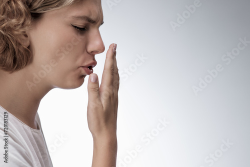 Bad breath. Young girl checking her breath with her hand, blowing to it, standing over grey background. Bad smell from the mouth, toothache, having problems with teeth